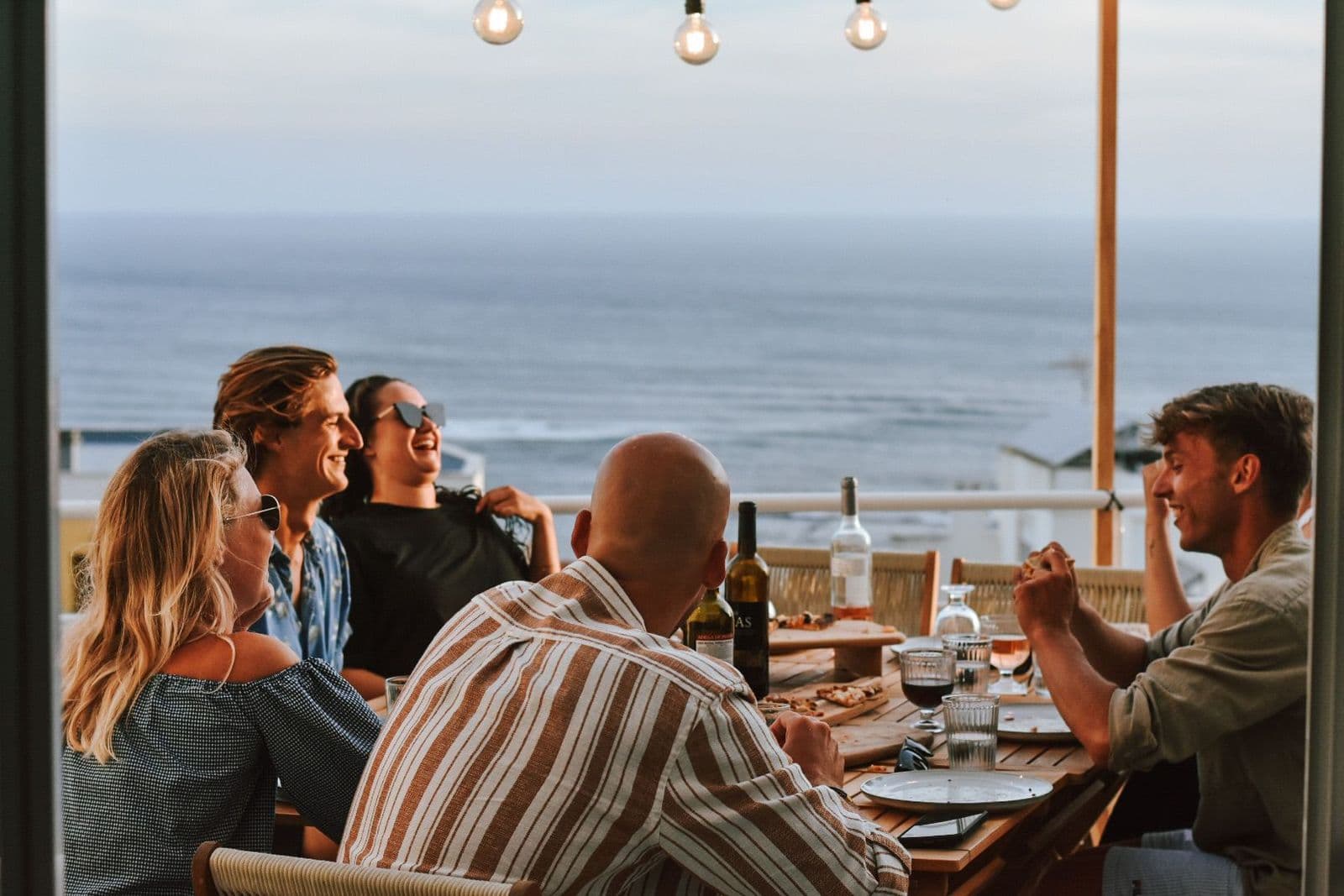 Vijf mensen zitten aan tafel te eten. Ze lachen en genieten van elkaars gezelschap. Op de achtergrond is de zee en er is een oranje gloed door de zonsondergang